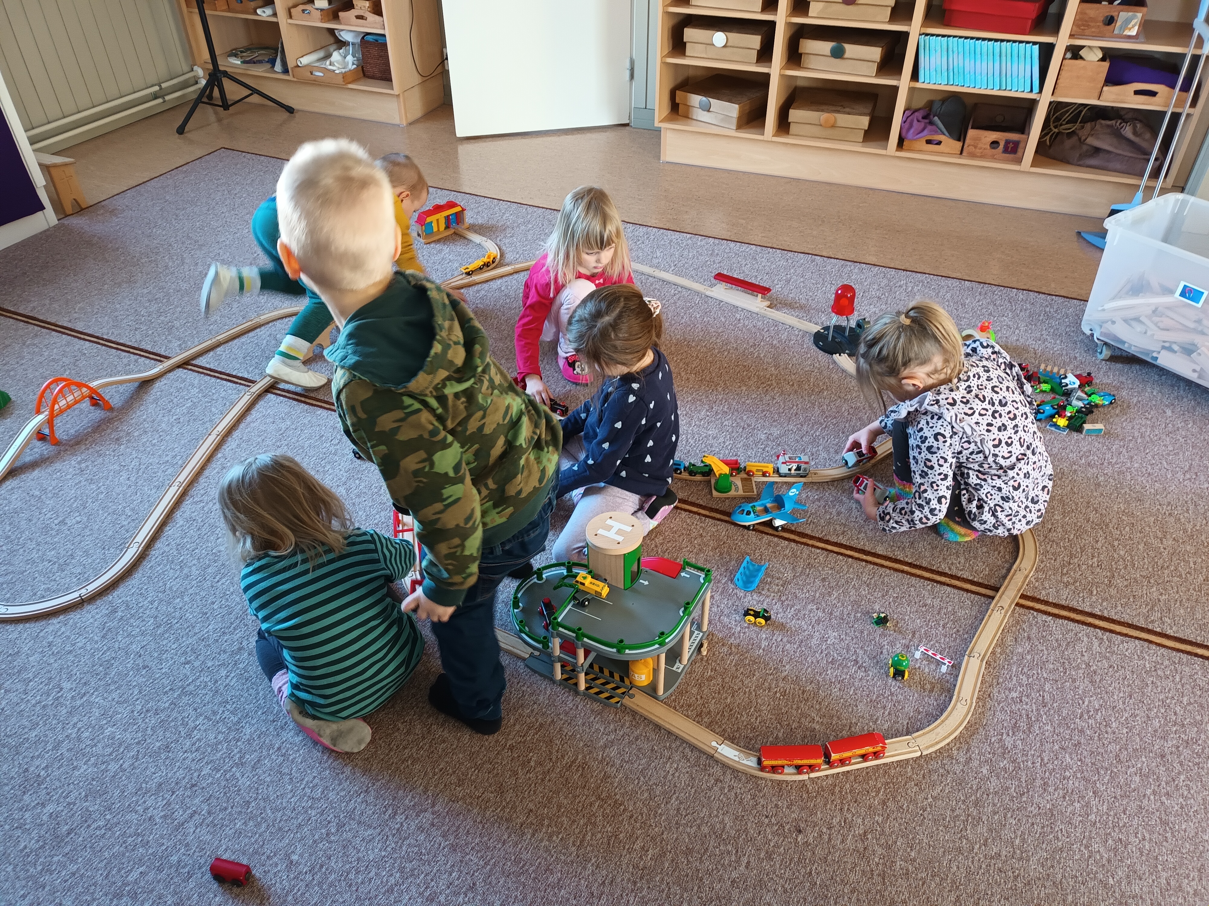 Kuusi päiväkerholaista leikkii yhteistä leikkiä Brio-junaradalla päiväkerhotilan lattialla.