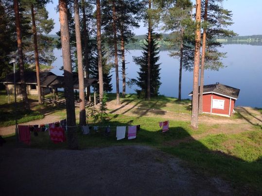 Peilityynen Sulkavajärven rannalla kylpee aamuauringossa saunamökki ja Anninkappeli. Pyykkinarulla kuivuu pyyhkeitä ja uimapukuja.