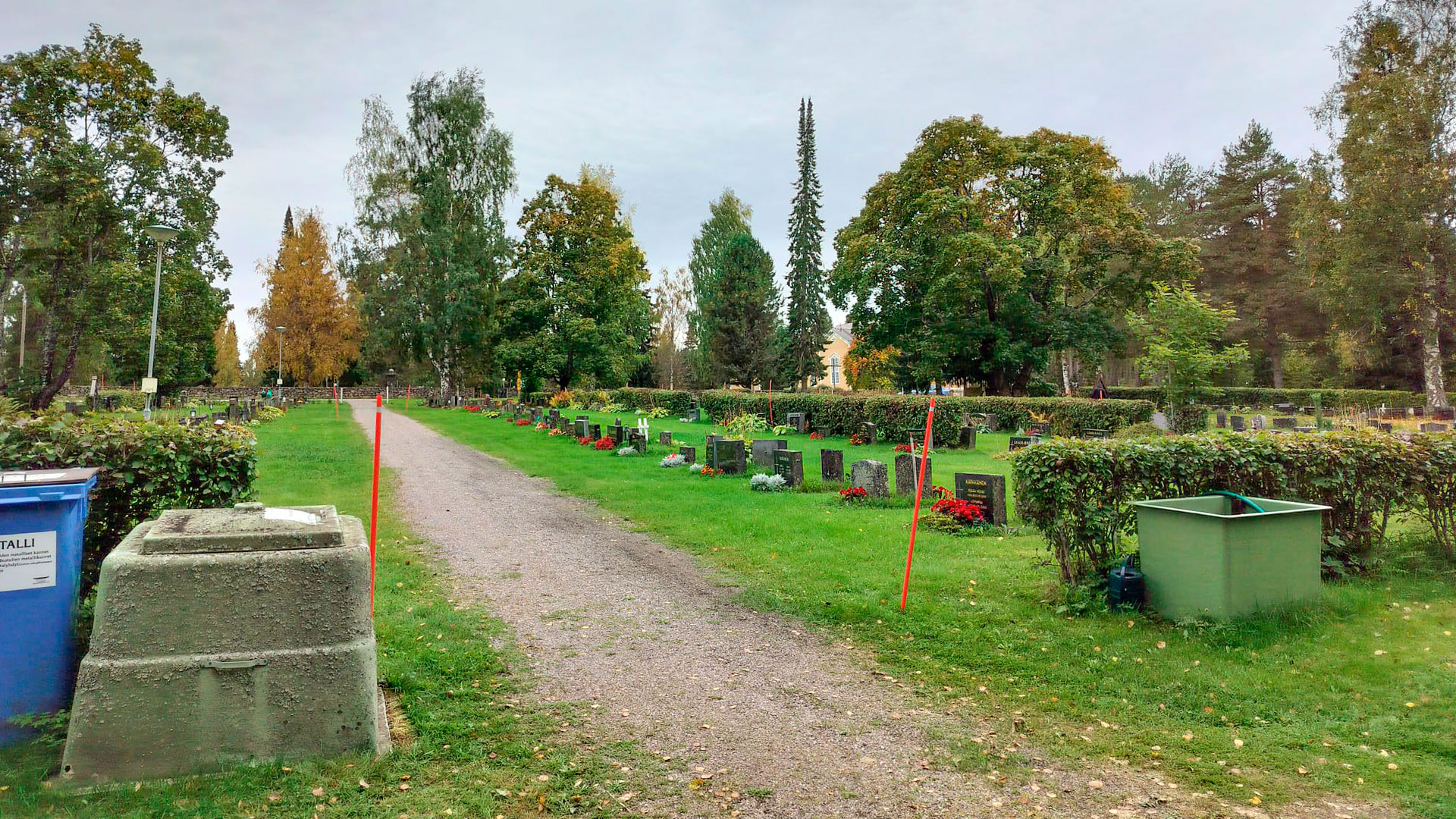 Hautausmaan hiekkakäytävä, hautakivia, nurmikkoa ja jäteastioita.