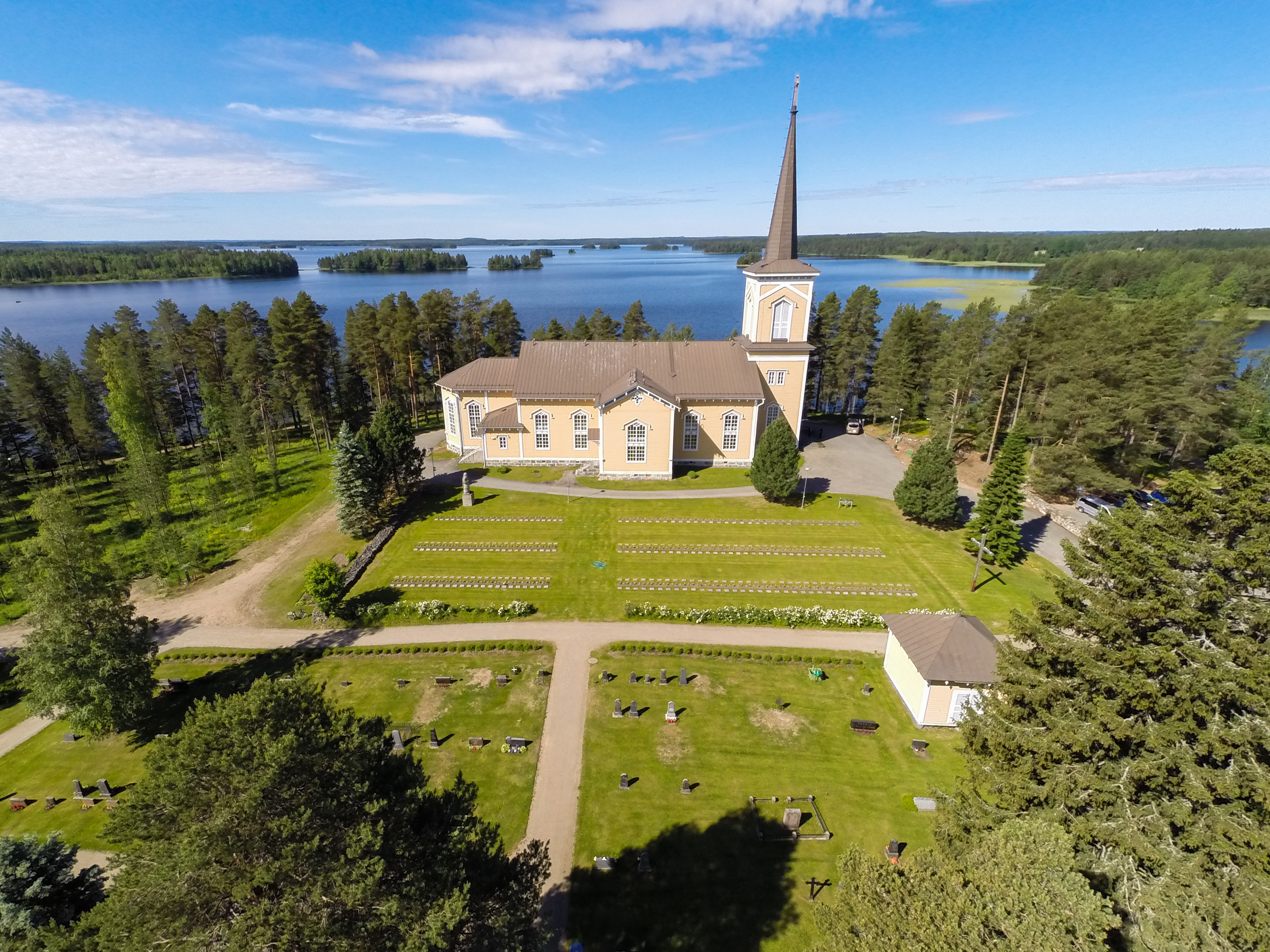 Ilmakuva Pielaveden kirkosta jossa näkyy kirkon lisäksi hautausmaata, järven rannalla suuria puita ja taust...