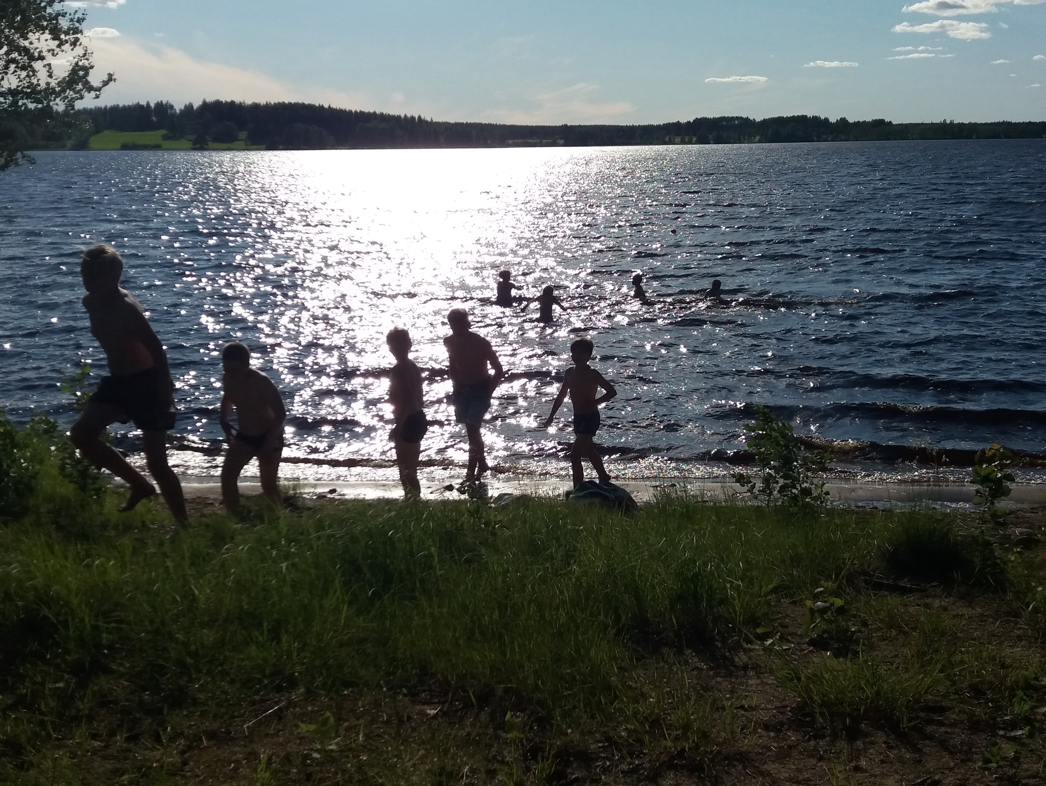 Lapsia menossa uimaan, aurinko kimmaltaa järven pinnasta.